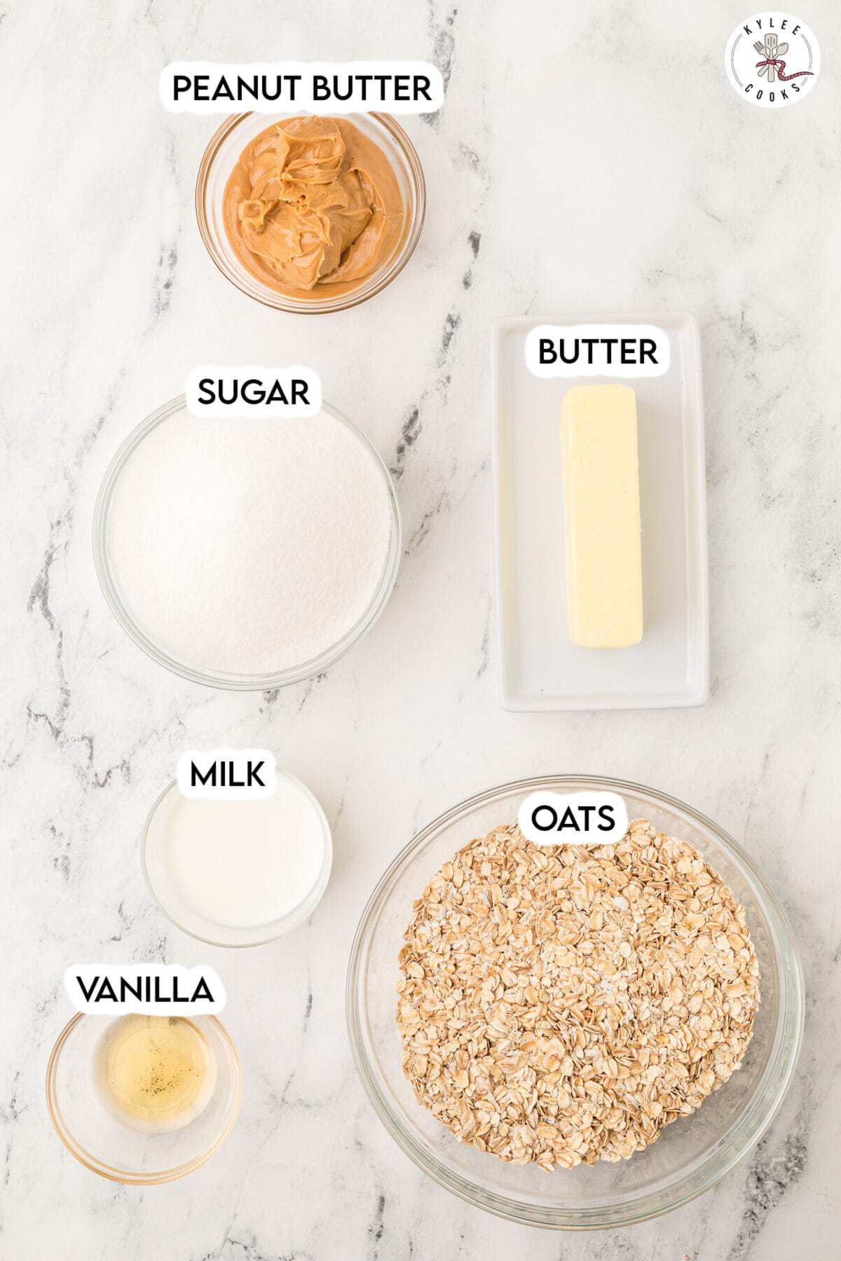 Five labeled ingredients on a marble countertop: peanut butter, sugar, butter, milk, vanilla, and oats, each in separate bowls or dishes.