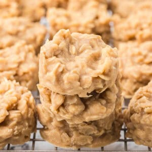 Three beige, round, no-bake cookies with oats stacked on a cooling rack, surrounded by more cookies in the background.
