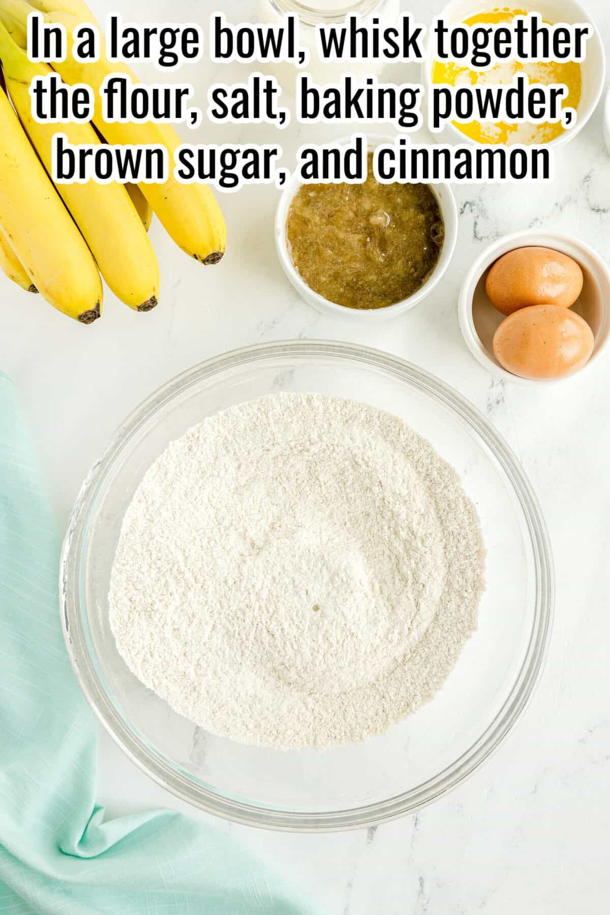 A glass bowl of flour mixture sits on a white counter near bananas, a bowl of mashed bananas, eggs, and a green cloth. Text with recipe instructions is overlaid at the top.