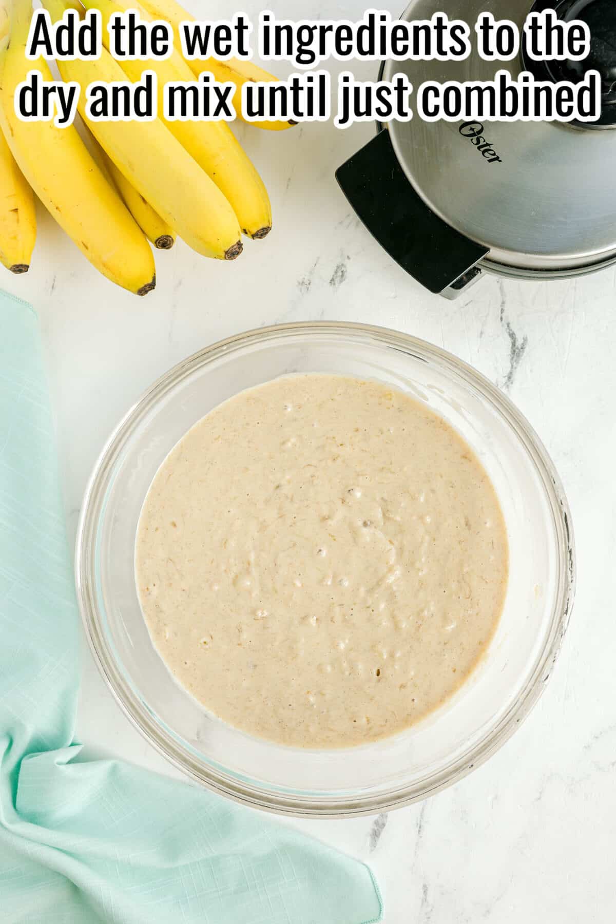 A glass bowl with banana bread batter sits on a white countertop beside bananas, a mixer, and a light blue cloth. Text above reads: “Add the wet ingredients to the dry and mix until just combined.”.