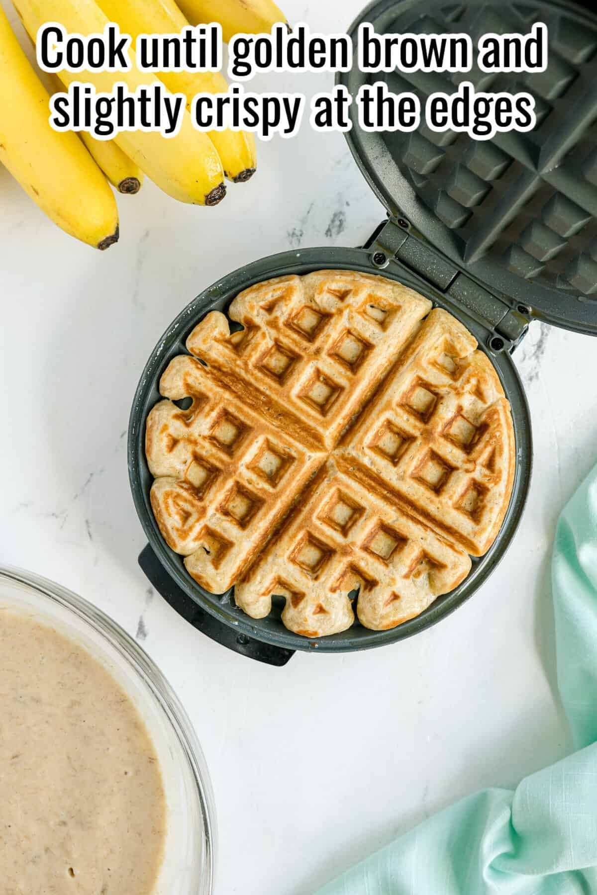 A round waffle in an open waffle maker sits on a marble surface near bananas, a bowl of batter, and a light blue cloth. Text at the top reads: "Cook until golden brown and slightly crispy at the edges.