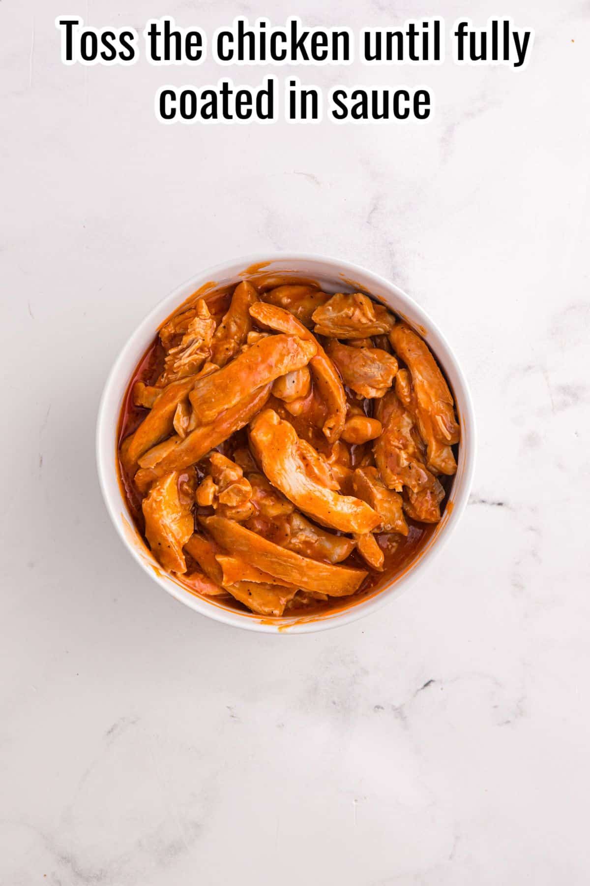 A bowl of cooked chicken strips coated in sauce sits on a white marble surface. Text above reads, "Toss the chicken until fully coated in sauce.