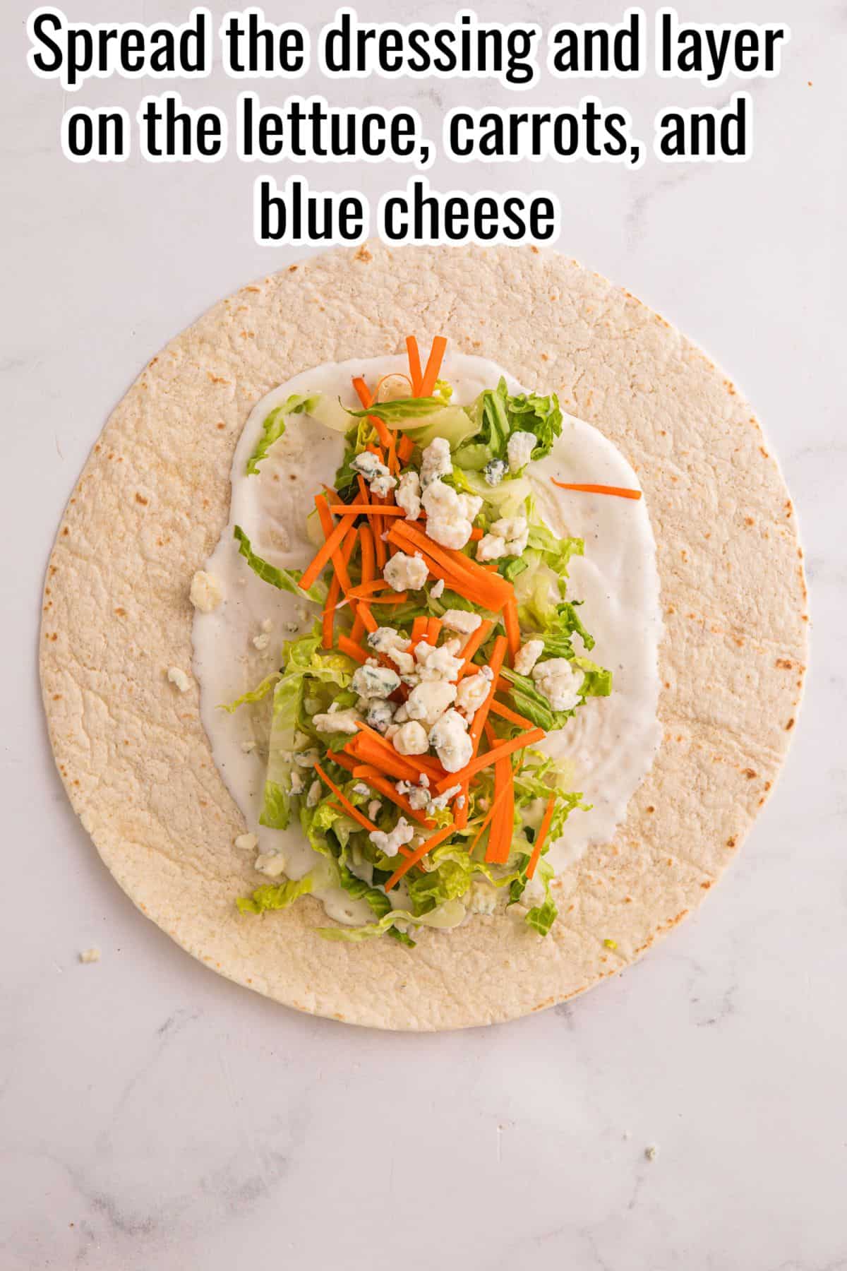 A tortilla topped with salad dressing, shredded lettuce, carrot strips, and crumbled blue cheese on a white surface.