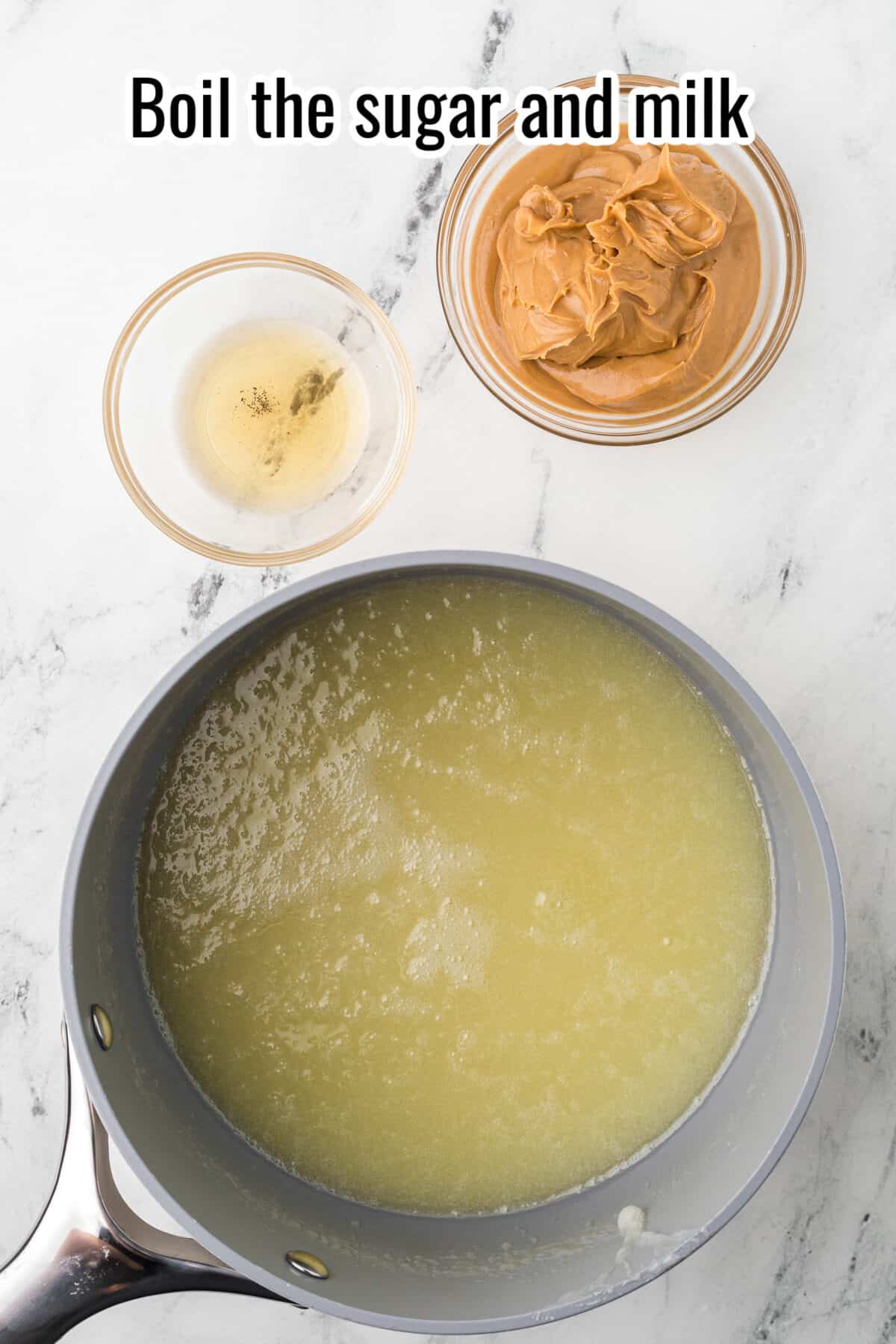 A saucepan with a boiling sugar and milk mixture, next to bowls of vanilla extract and peanut butter, on a marble countertop. Text above reads, "Boil the sugar and milk.