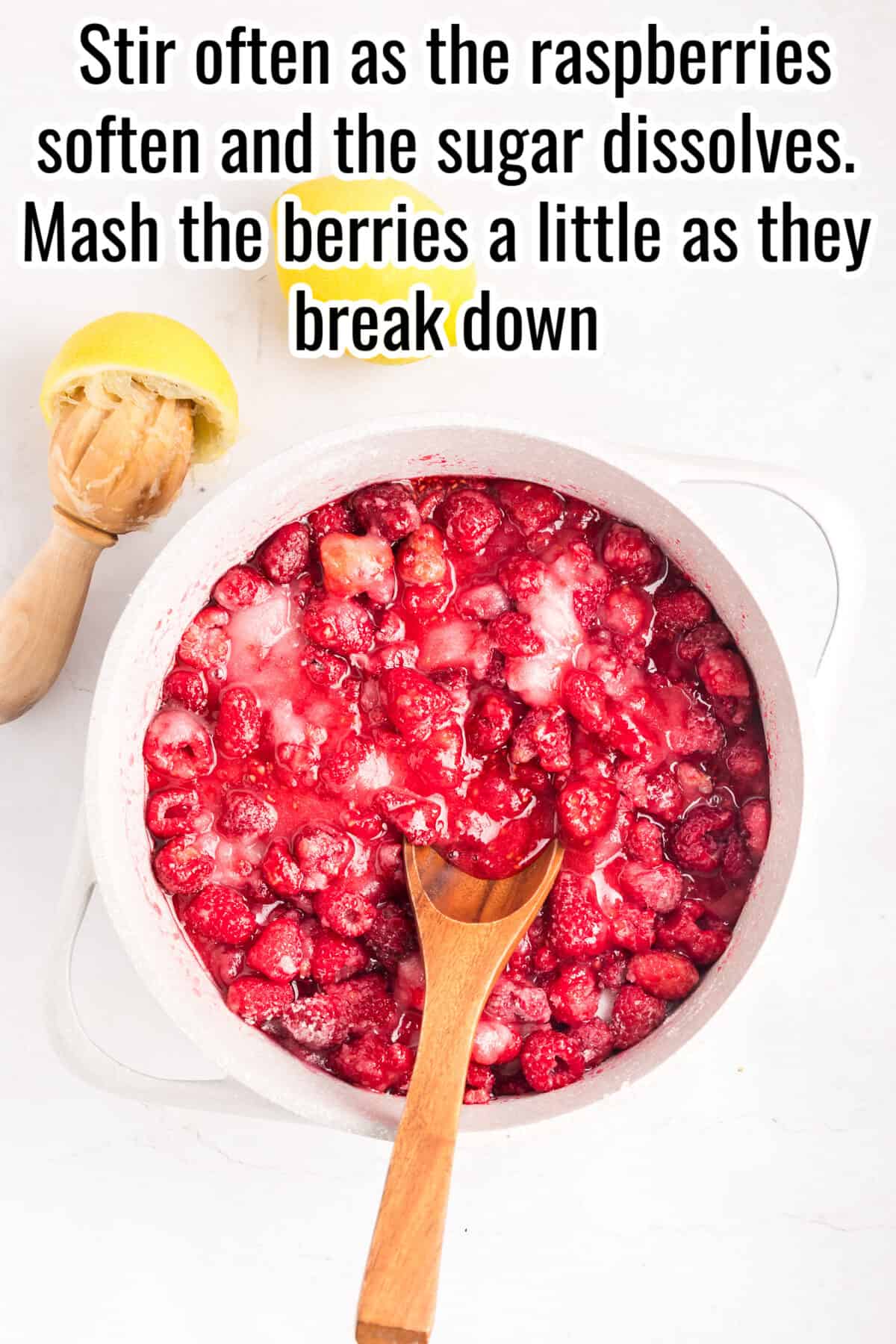 A white pot of mashed raspberries with a wooden spoon sits on a white surface next to a lemon squeezer and a halved lemon. Text instructions are above the pot.