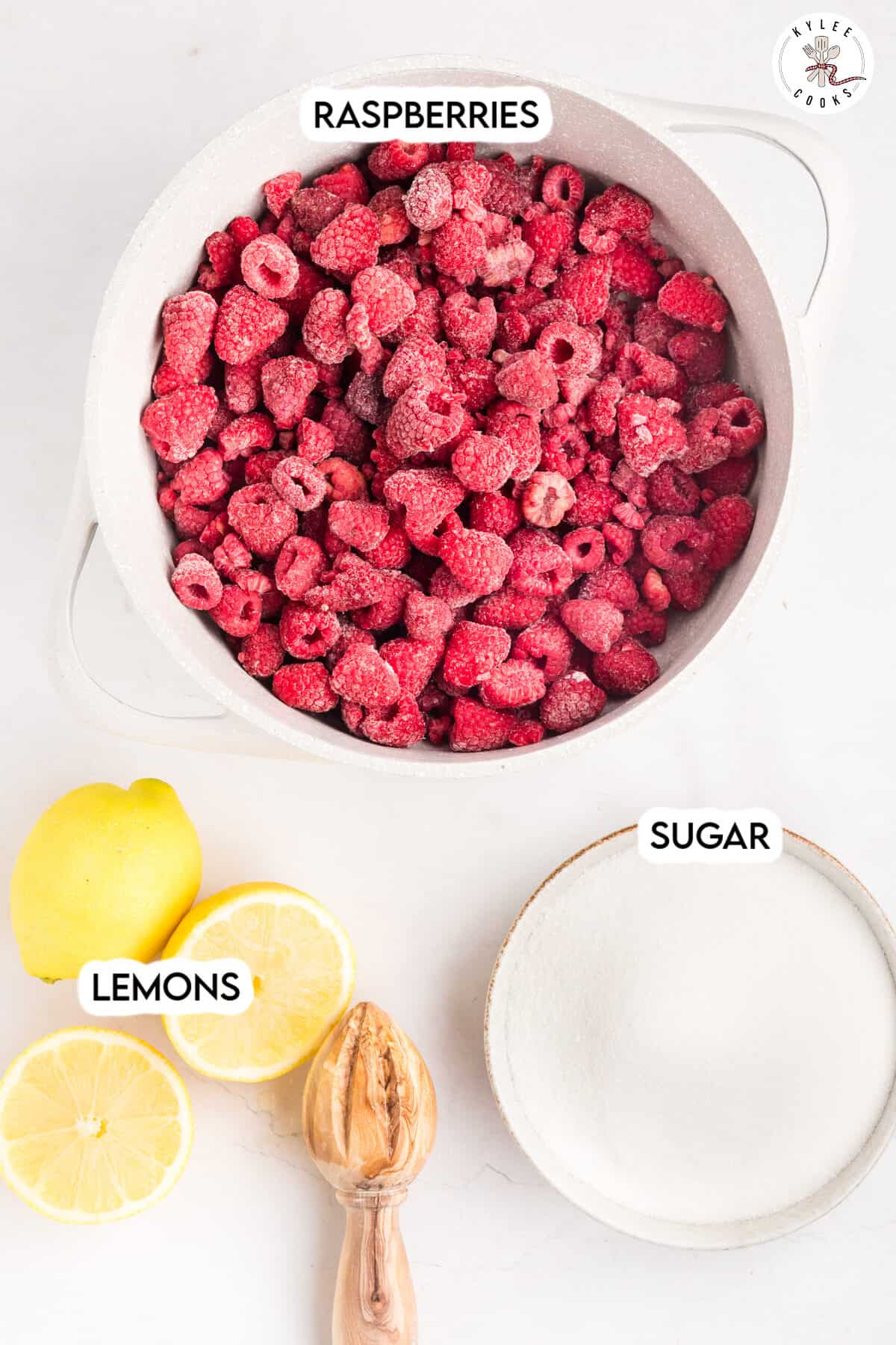 A pot of raspberries, a bowl of sugar, whole and halved lemons, and a wooden lemon juicer are arranged on a white surface.