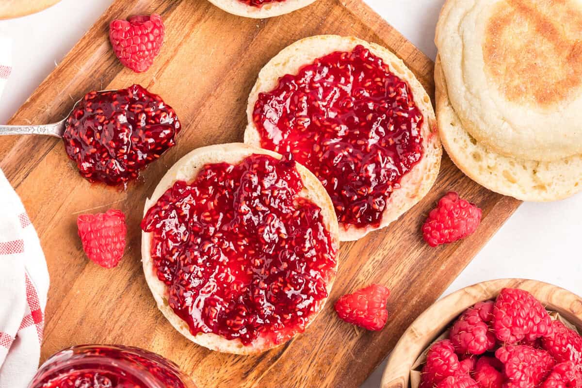 Two English muffins with raspberry jam on a wooden board, surrounded by fresh raspberries and a spoonful of jam.