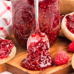 A spoon with raspberry jam in front of a mason jar, surrounded by pieces of bread spread with jam and fresh raspberries on a wooden surface.