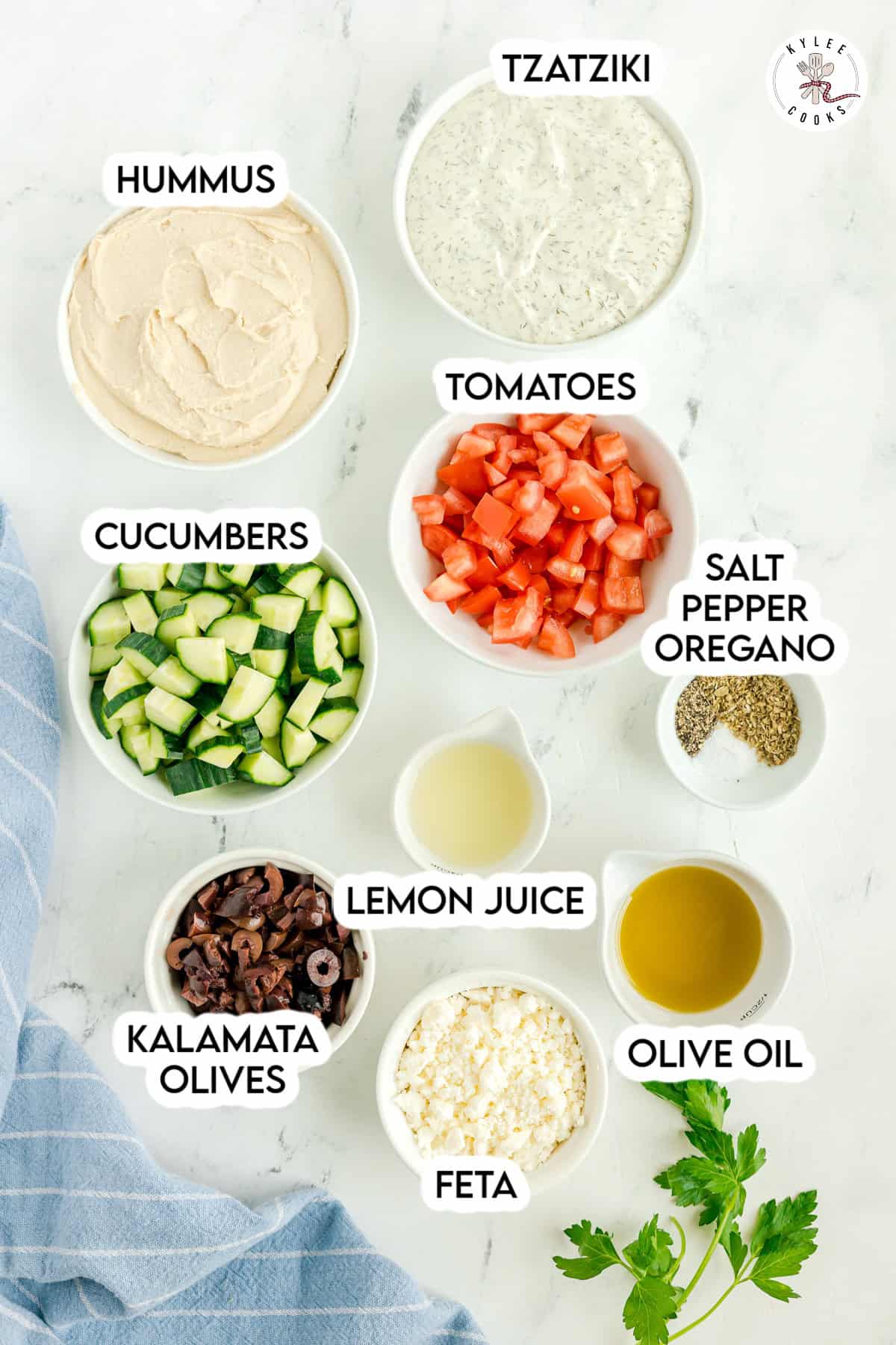 Bowls containing hummus, tzatziki, chopped tomatoes, cucumbers, kalamata olives, feta cheese, lemon juice, olive oil, and a seasoning mix are arranged on a white surface—perfect ingredients for creating a vibrant Mediterranean dip or a classic Layered Greek Dip.