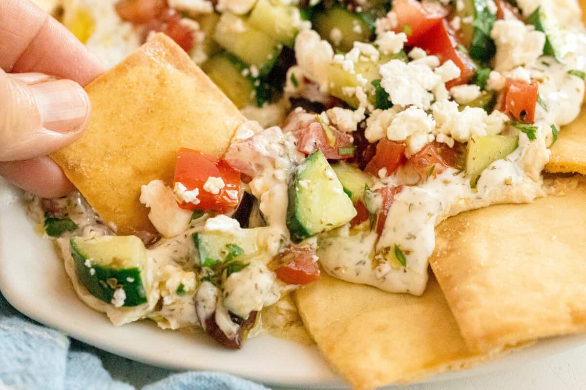 Hand dipping pita chip into Mediterranean-style dip with chopped tomatoes, cucumbers, olives, and crumbled feta cheese on a white plate.