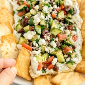 A hand dips pita chip into Greek layer dip topped with chopped cucumber, tomato, olives, and feta cheese, surrounded by pita chips on a white plate.