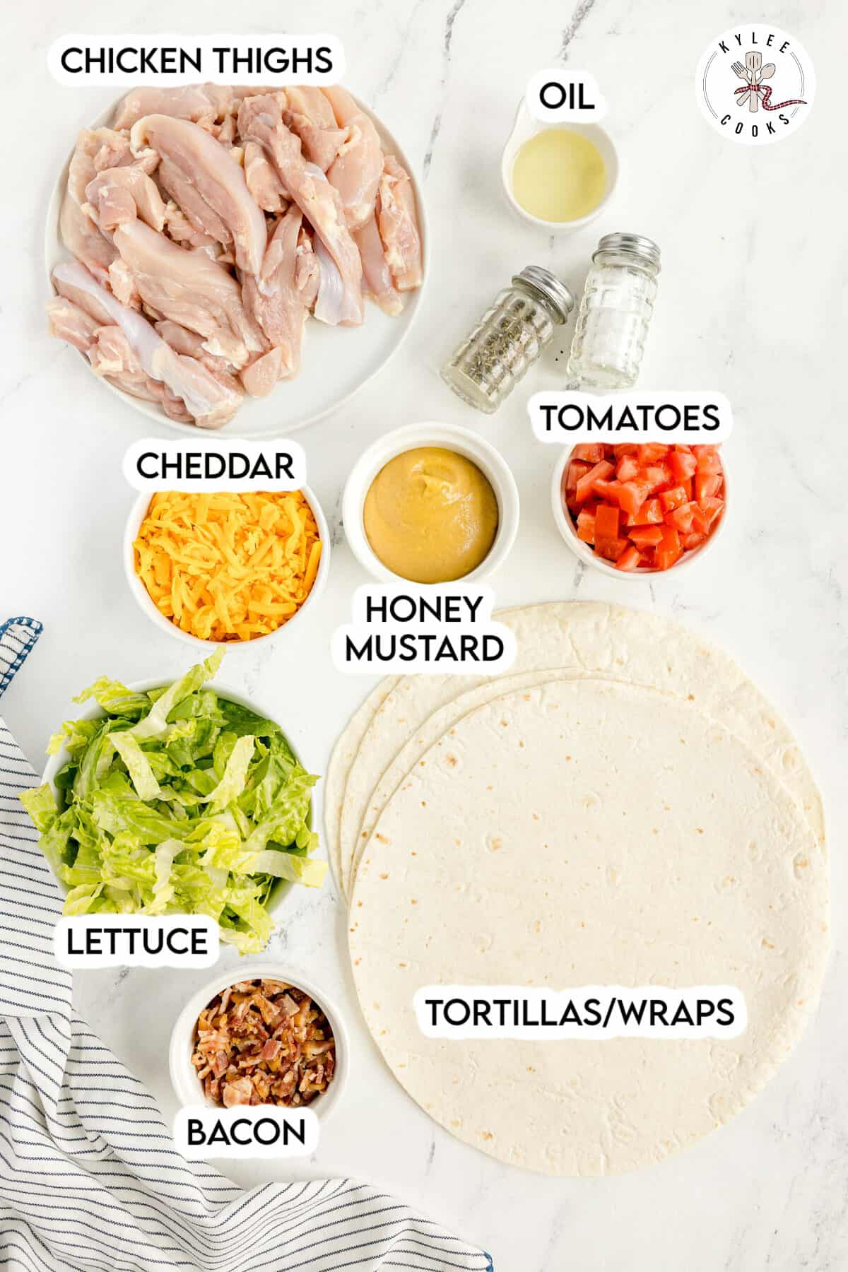 Ingredients for wraps are arranged on a counter: chicken thighs, oil, cheddar cheese, tomatoes, honey mustard, lettuce, bacon, and tortillas.