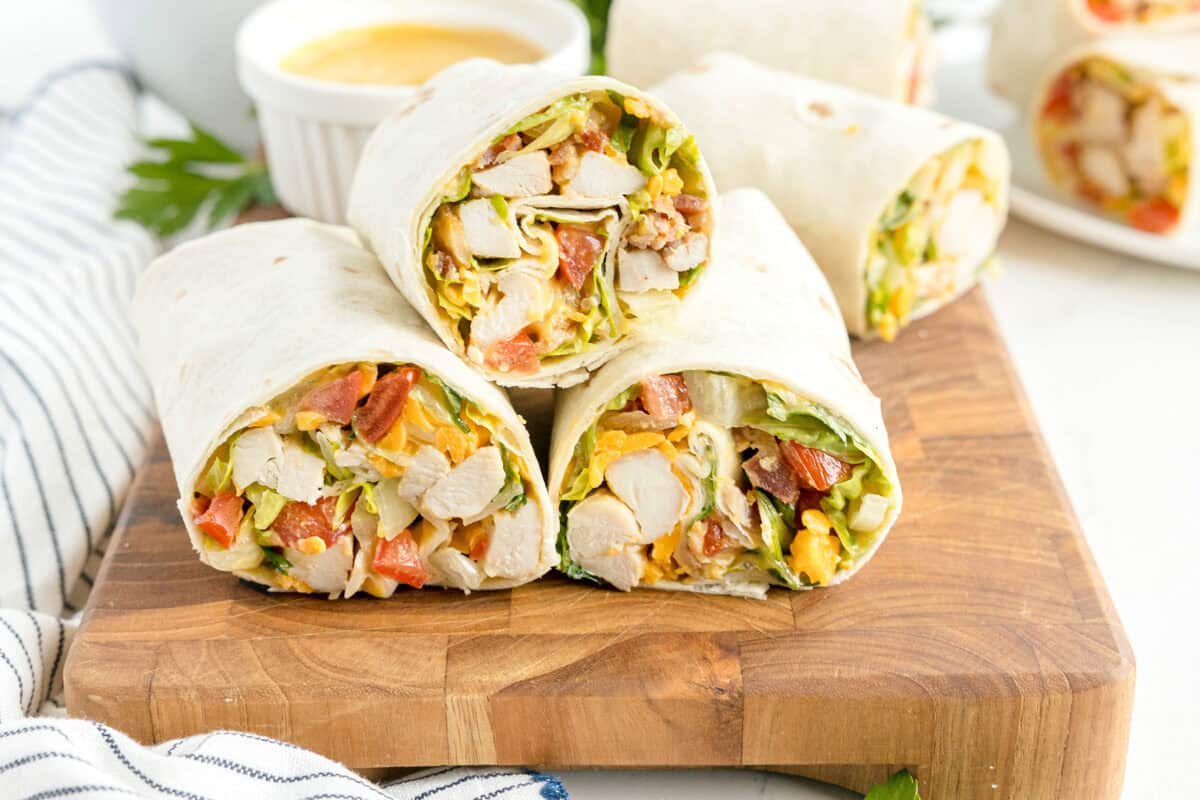 Three chicken wraps with lettuce, tomato, and cheese are stacked on a wooden board. A small cup of sauce and more wraps are in the background.