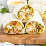 Three chicken wraps filled with lettuce, tomato, and cheese are stacked on a wooden board, with a cup of yellow dipping sauce in the background.