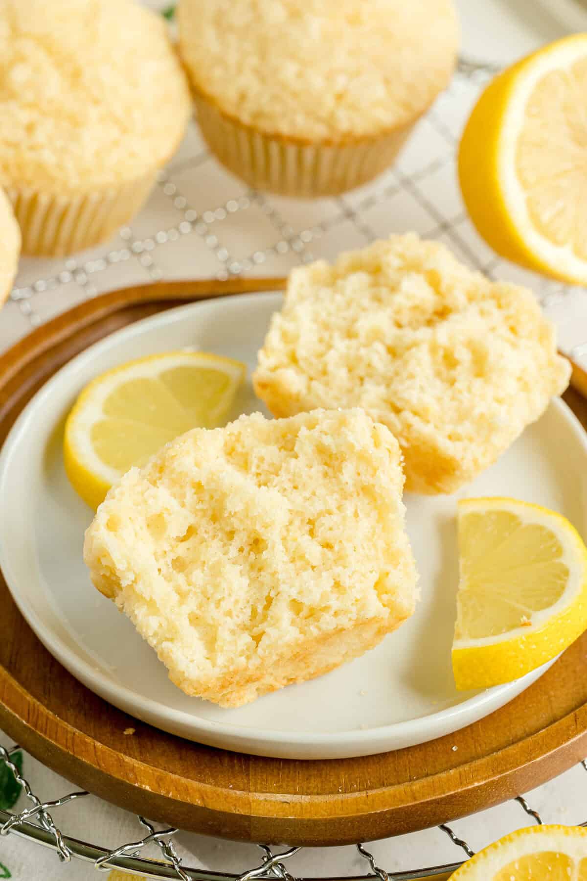 A split lemon muffin sits on a white plate with two lemon wedges, with more muffins and lemon halves in the background.