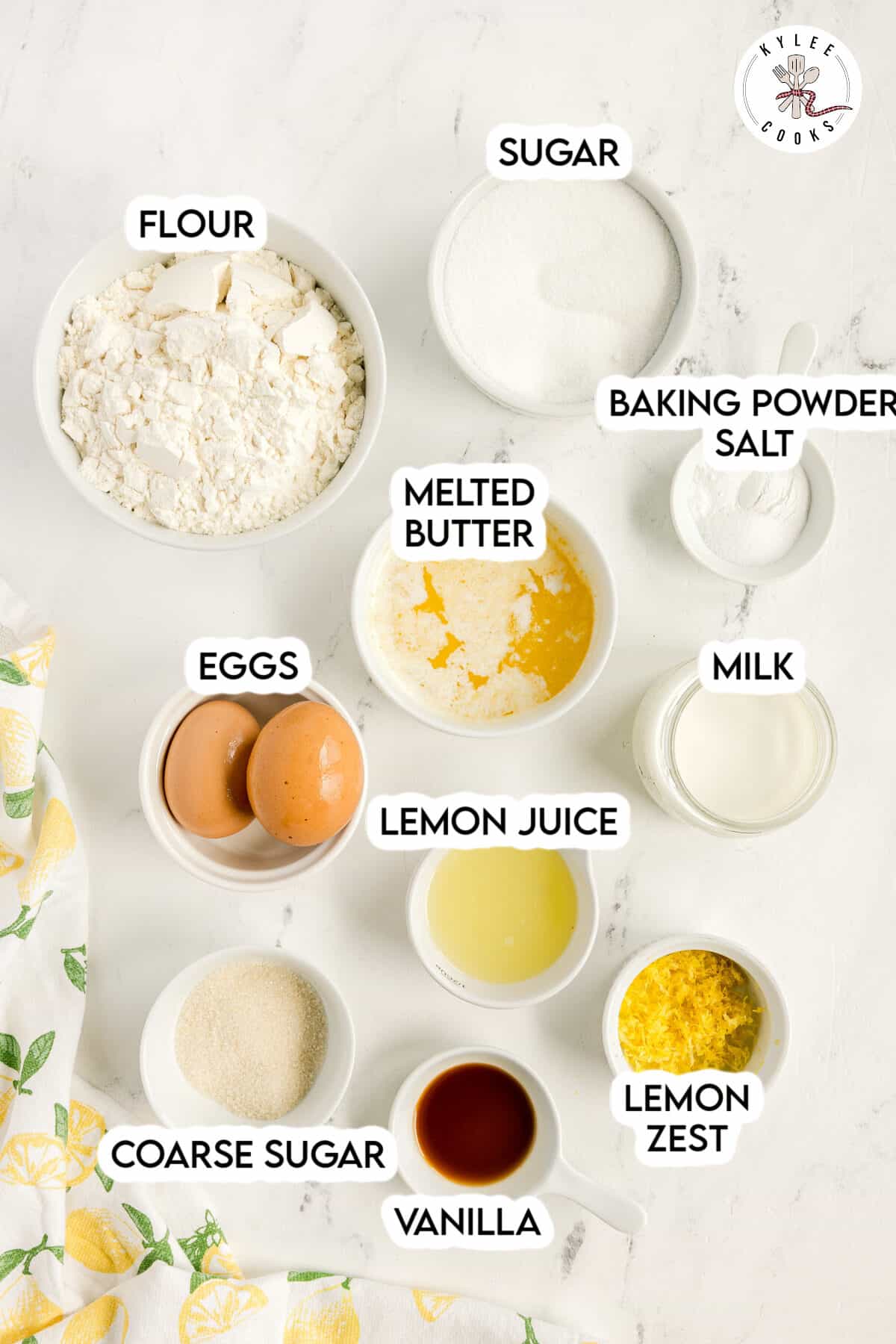 Baking ingredients on a white surface, labeled: flour, sugar, baking powder, salt, milk, eggs, melted butter, lemon juice, lemon zest, coarse sugar, and vanilla.