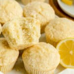 Close-up of several lemon muffins, one with a bite taken out, with a lemon slice nearby on a white surface.