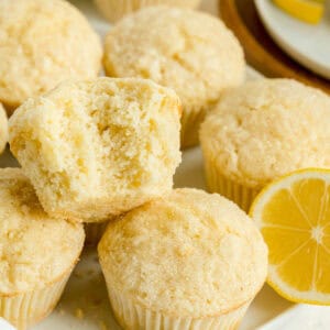 Close-up of several lemon muffins, one with a bite taken out, with a lemon slice nearby on a white surface.