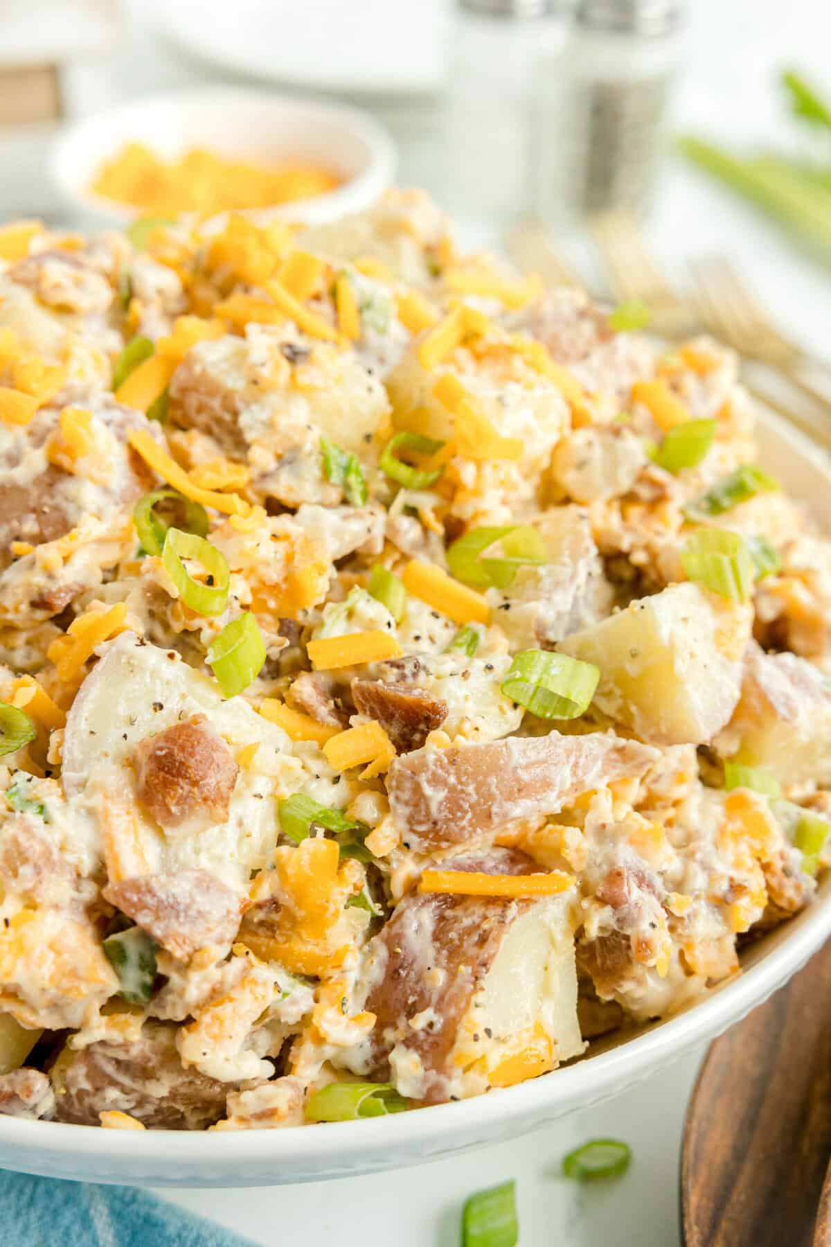 A close-up of a bowl filled with creamy potato salad topped with shredded cheddar cheese and chopped green onions.