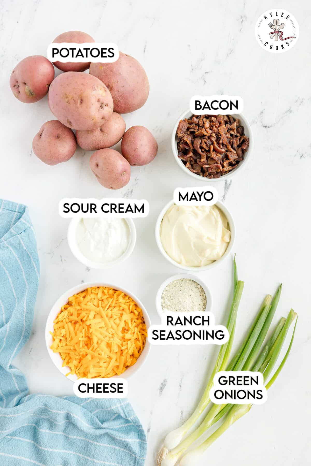 Six whole potatoes, a bowl of bacon, sour cream, mayo, shredded cheese, ranch seasoning, and three green onions arranged on a white surface with a blue towel.