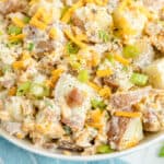 A close-up of a bowl of potato salad with diced potatoes, shredded cheddar cheese, bacon bits, sliced green onions, and creamy dressing.