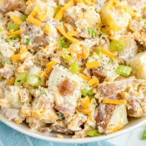 A close-up of a bowl of potato salad with diced potatoes, shredded cheddar cheese, bacon bits, sliced green onions, and creamy dressing.