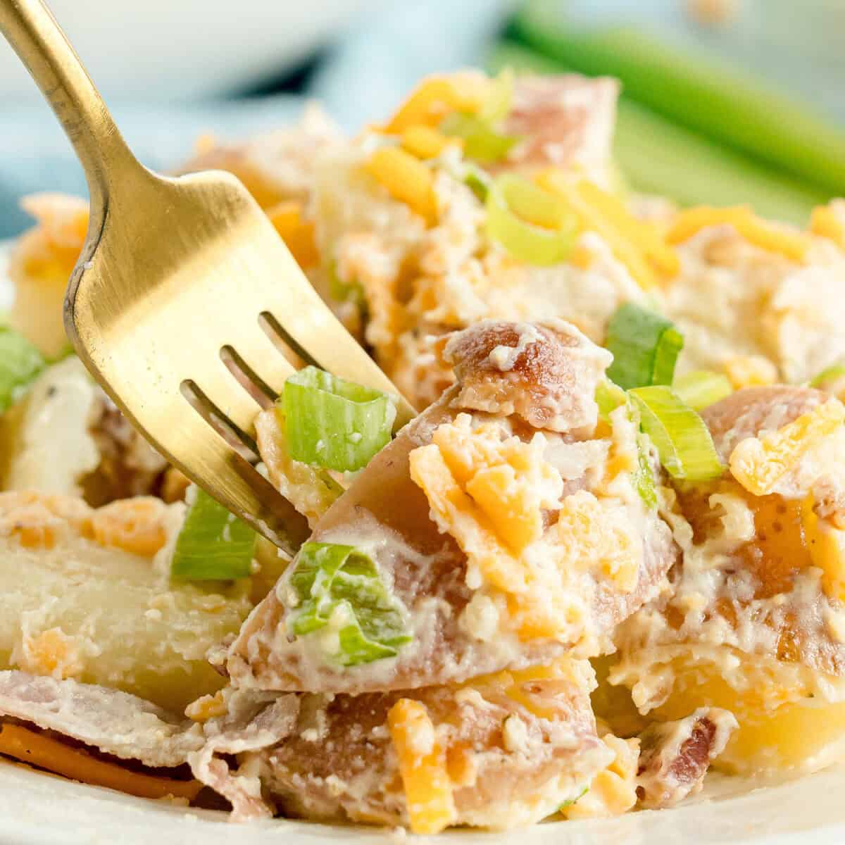 A fork picking up a bite of potato salad with shredded cheddar cheese, chopped green onions, and creamy dressing.