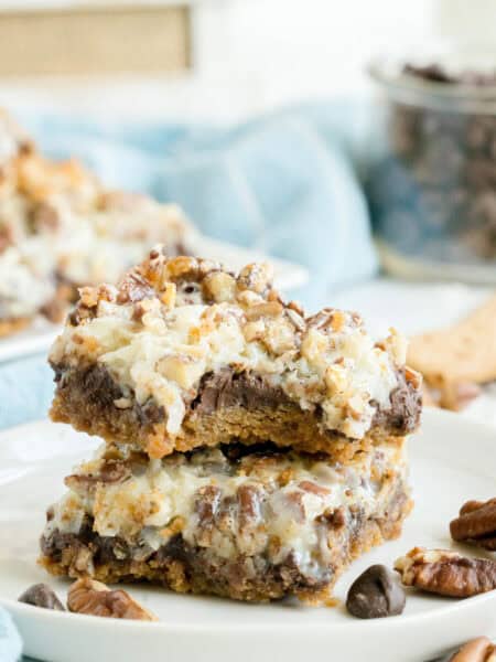 Two stacked dessert bars with chocolate chips, nuts, and a gooey topping sit on a white plate, with more dessert bars and ingredients visible in the blurred background.