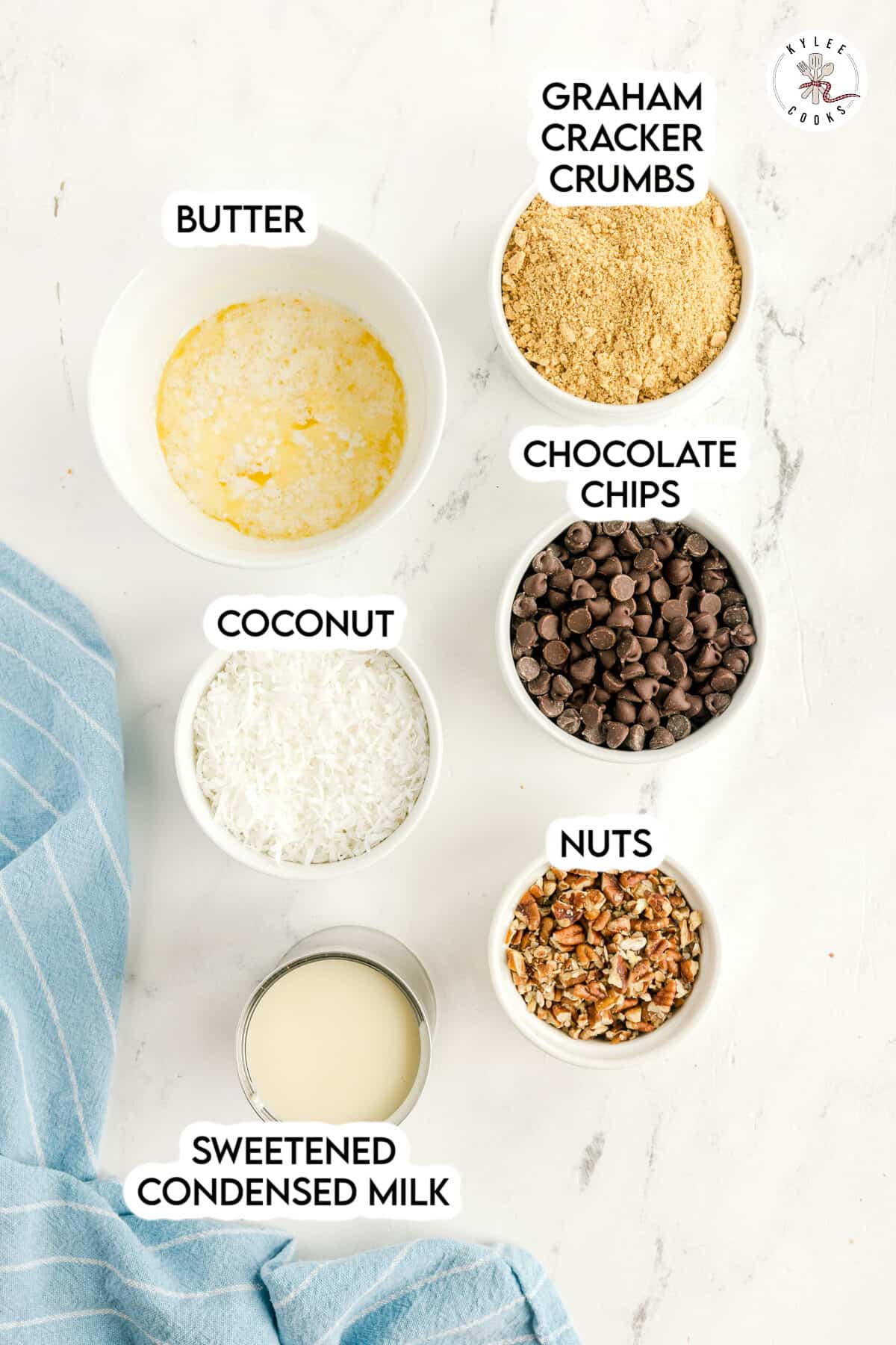 Six labeled bowls and cups containing butter, graham cracker crumbs, chocolate chips, coconut, nuts, and sweetened condensed milk are arranged on a white surface next to a blue cloth.