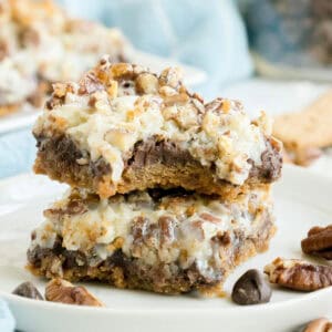 Two dessert bars with a crumbly base, chocolate chips, chopped nuts, and a creamy topping are stacked on a white plate, with extra nuts and chocolate chips beside them.