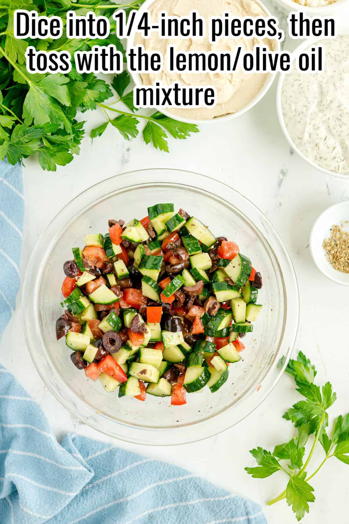 A glass bowl of chopped cucumber, tomato, and olives sits on a white surface with fresh herbs, spices, and a towel nearby. Text reads: “Dice into 1/4-inch pieces, then toss with the lemon/olive oil mixture.”.