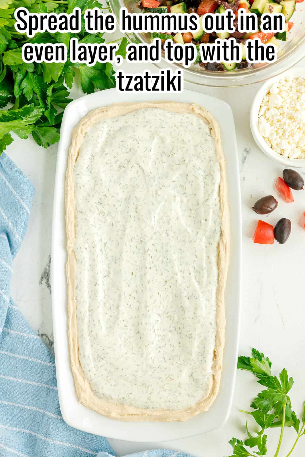 A rectangular dish with a layer of hummus topped with tzatziki, surrounded by parsley, olives, feta, and a blue striped cloth.