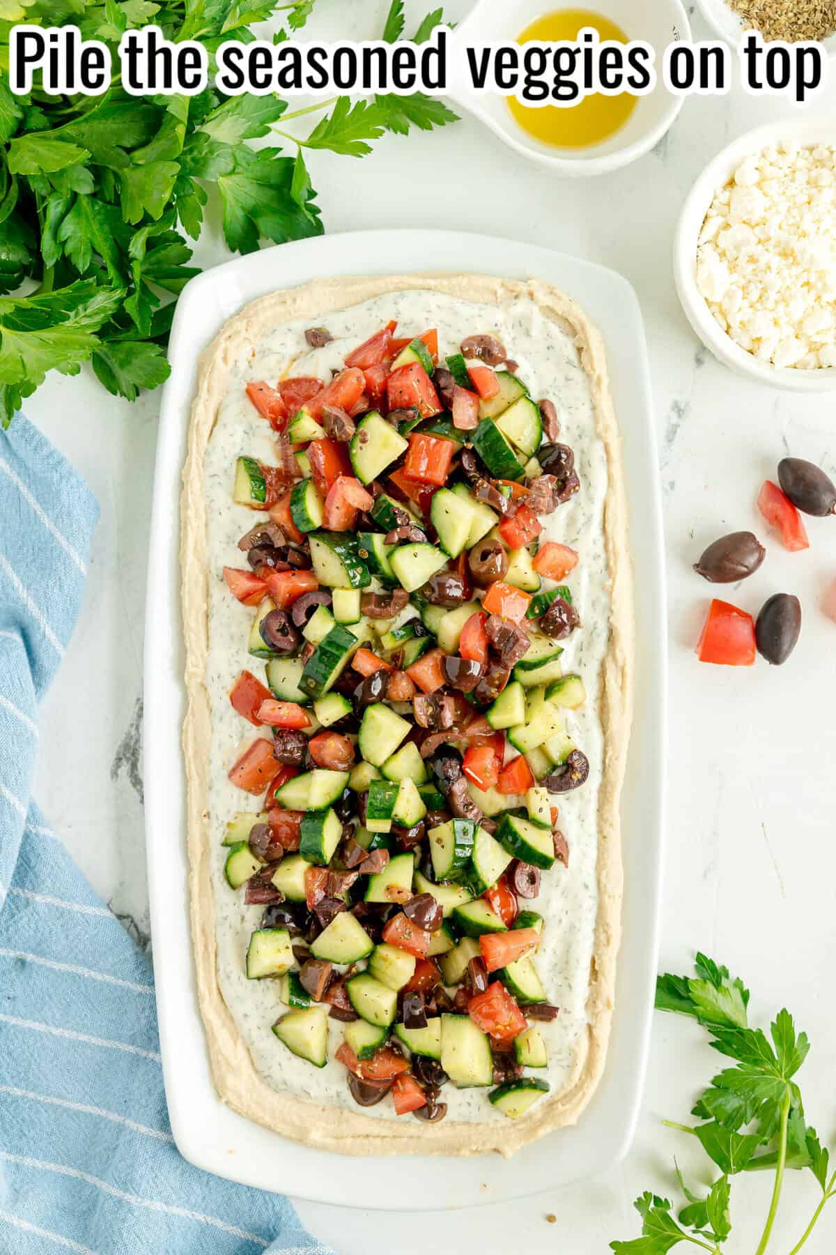 A rectangular platter with a hummus spread topped with chopped cucumbers, tomatoes, and olives, surrounded by parsley, feta, olives, and olive oil on a white surface.
