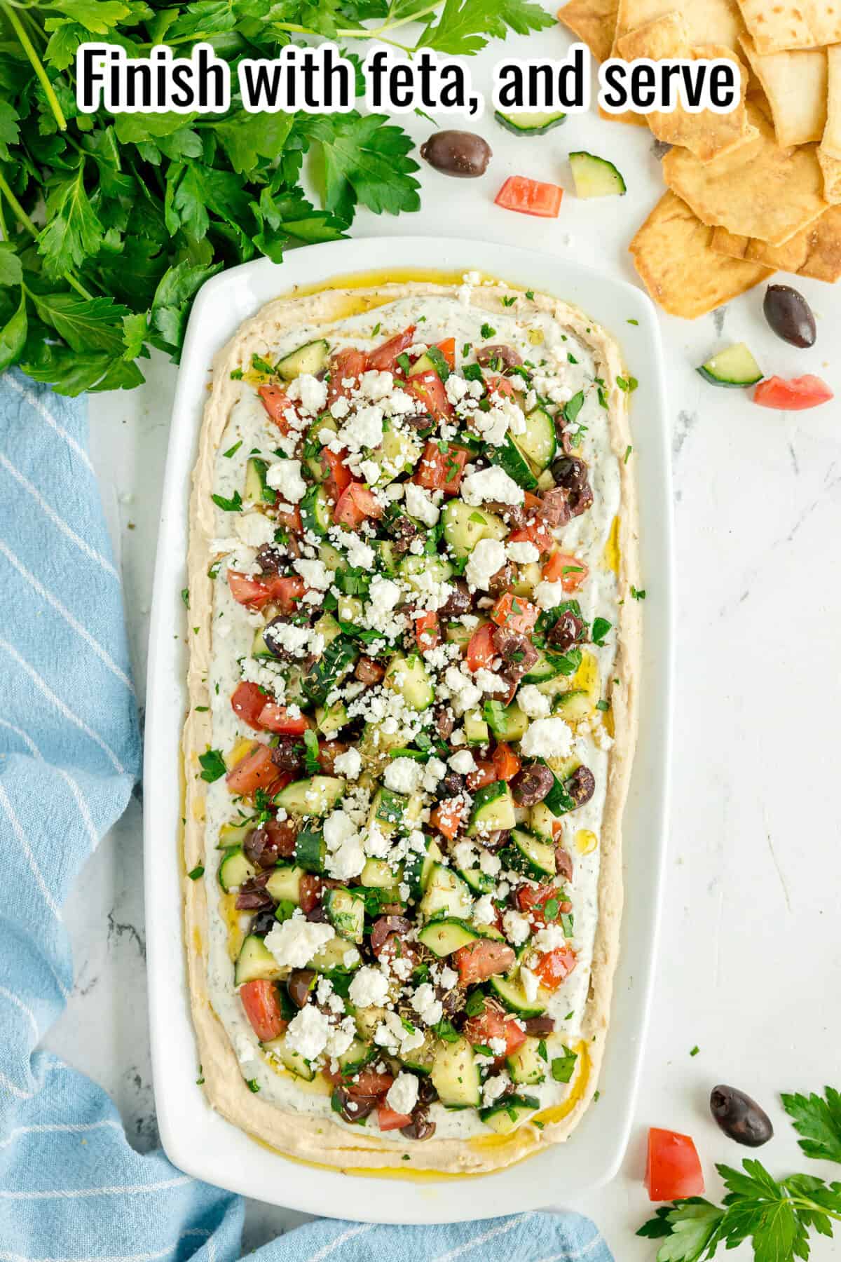 Rectangular platter of Mediterranean dip topped with chopped cucumbers, tomatoes, olives, feta, and herbs, surrounded by pita chips and fresh parsley. Text reads: “Finish with feta, and serve.”.