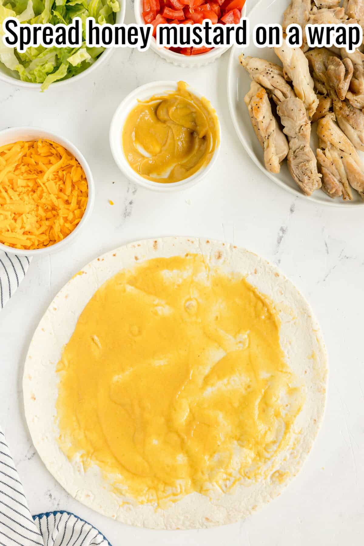 A tortilla wrap spread with honey mustard, surrounded by bowls of cheddar cheese, lettuce, tomatoes, and cooked chicken strips.