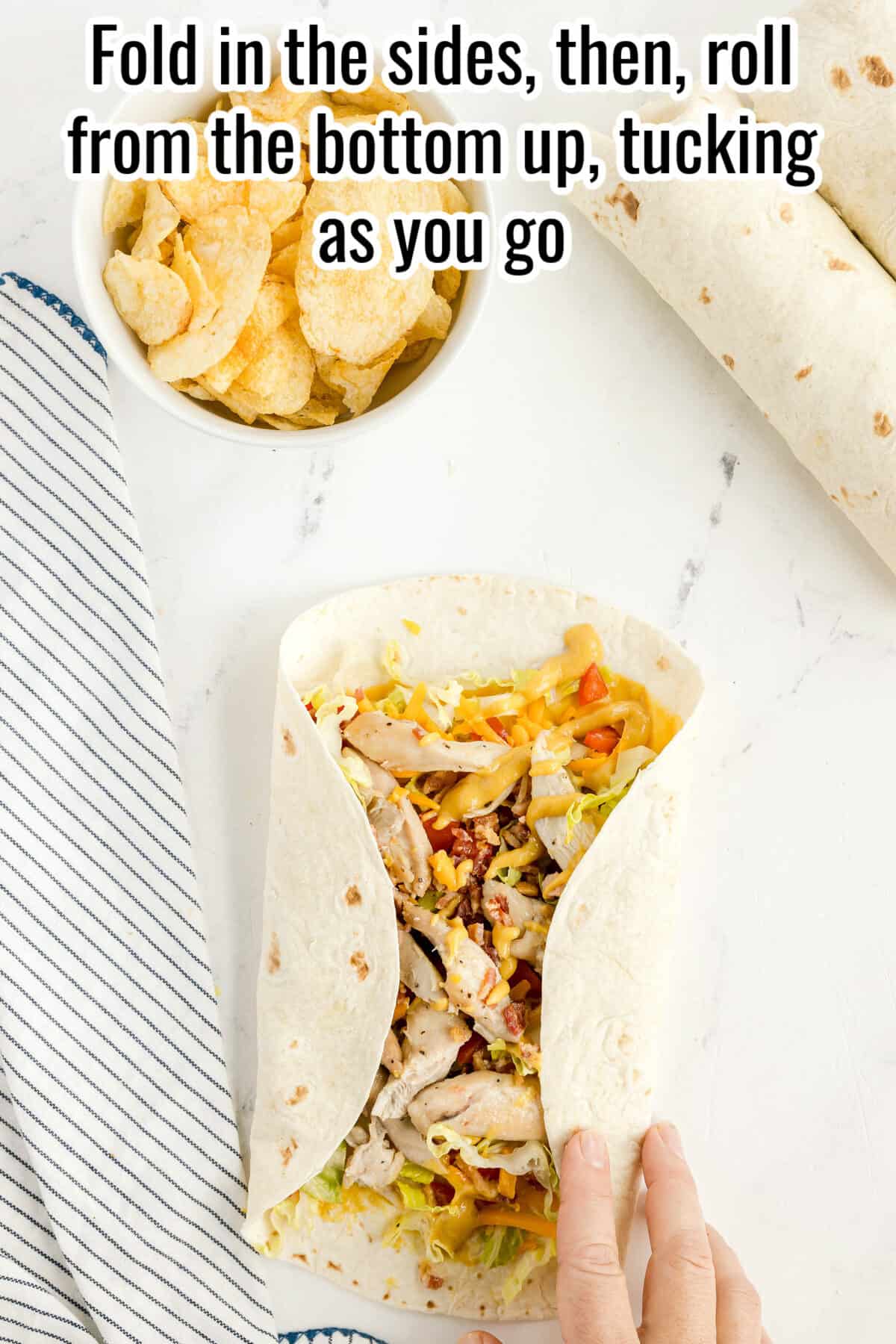 A hand folds a tortilla wrap filled with chicken and vegetables on a white surface, with a bowl of chips and a striped napkin nearby. Text at the top gives rolling instructions.