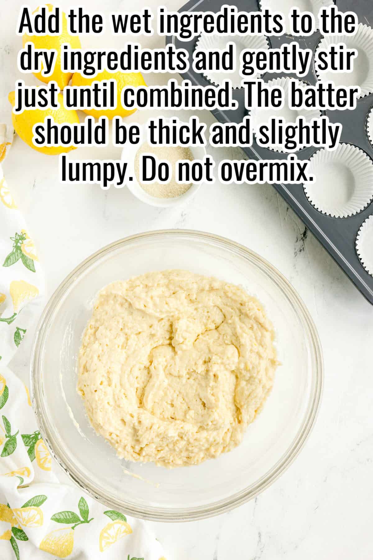 A glass bowl with thick, slightly lumpy batter on a white surface, next to a muffin tin with paper liners and a tea towel with lemon graphics.