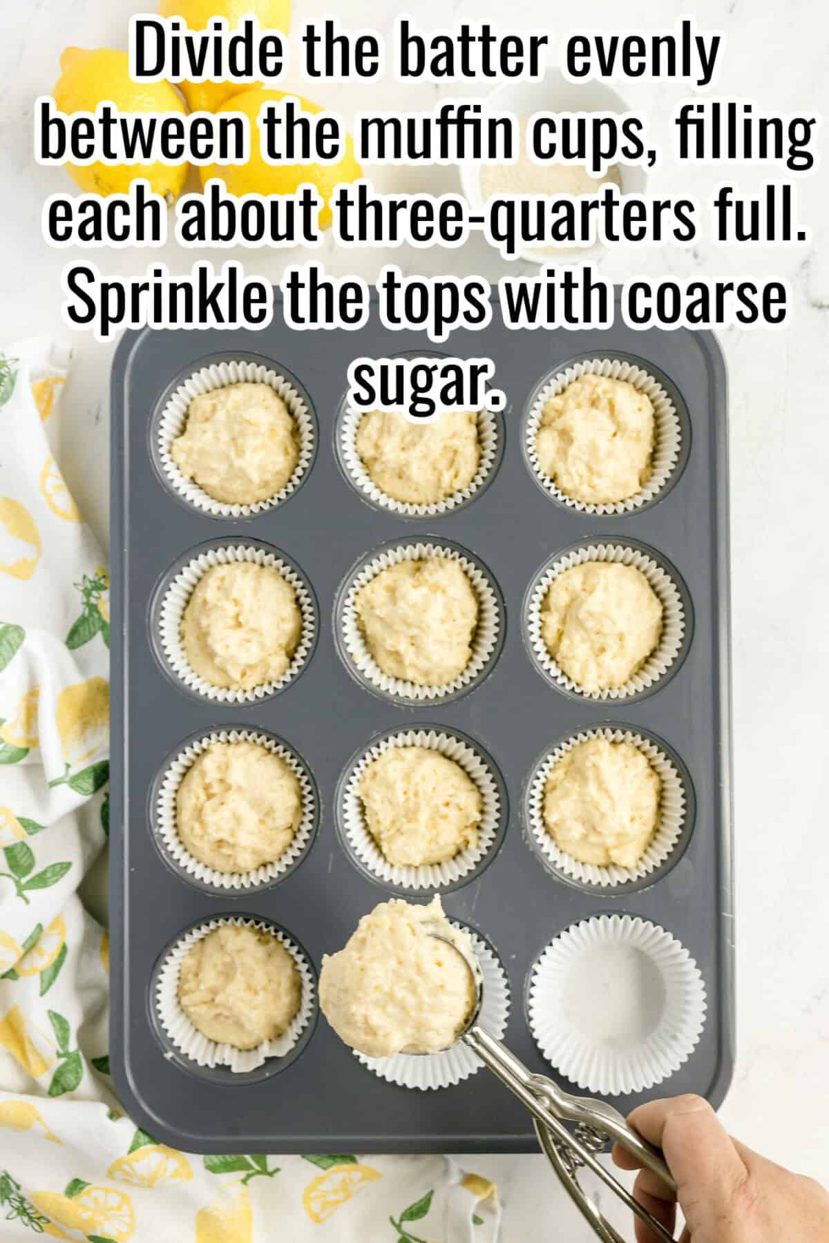 A hand uses an ice cream scoop to fill a lined muffin tin with batter. Most cups are filled about three-quarters full. Text instructions are overlaid at the top.
