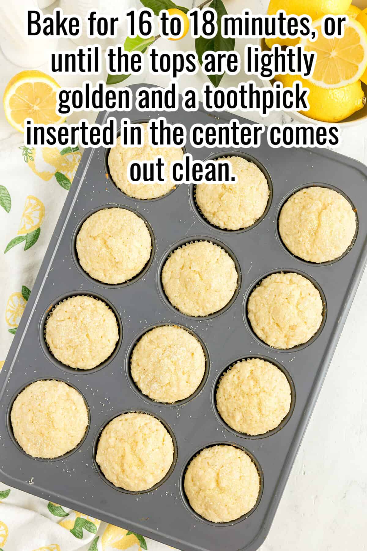 A muffin tin filled with twelve baked muffins sits on a light surface with lemon-patterned cloth and lemon slices nearby. Text with baking instructions is overlaid at the top of the image.