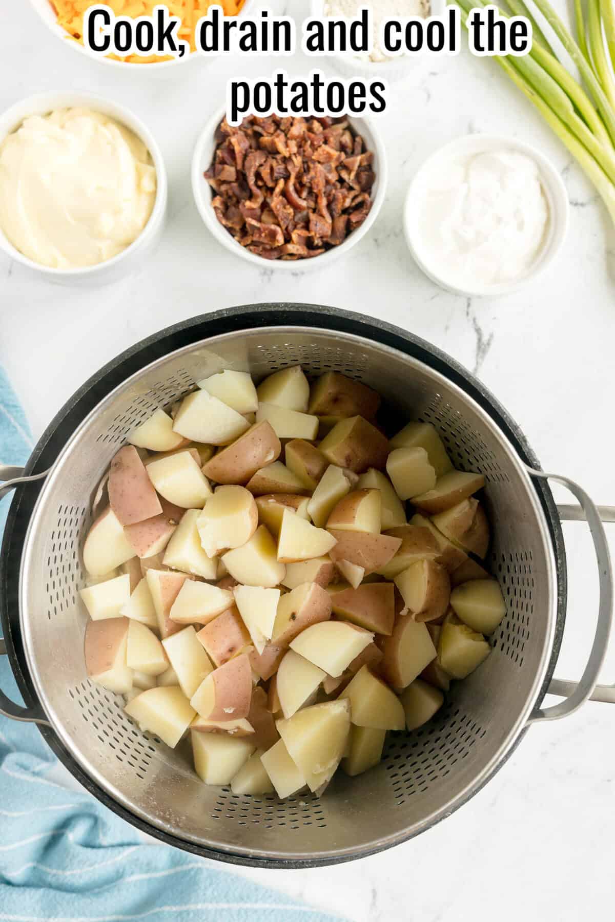 A colander filled with cooked, chopped potatoes sits on a counter next to bowls of mayonnaise, chopped bacon, sour cream, and green onions. Text reads: “Cook, drain and cool the potatoes.”.