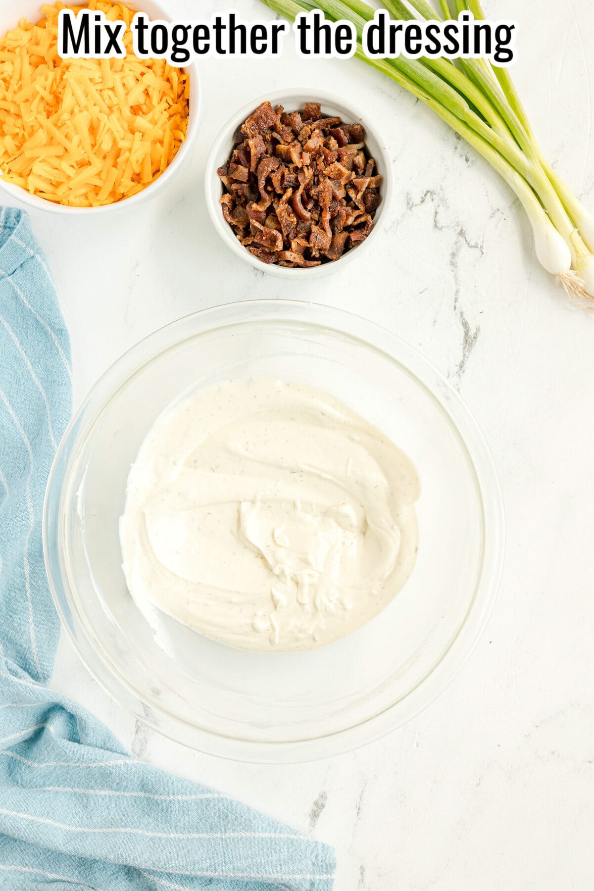 A glass bowl of creamy dressing sits on a white surface, surrounded by shredded cheese, chopped bacon, green onions, and a blue towel. Text at top reads, “Mix together the dressing.”.