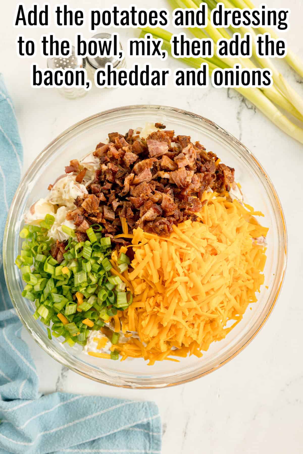 A glass bowl with chopped bacon, shredded cheddar cheese, sliced green onions, and potato salad ingredients on a white surface.