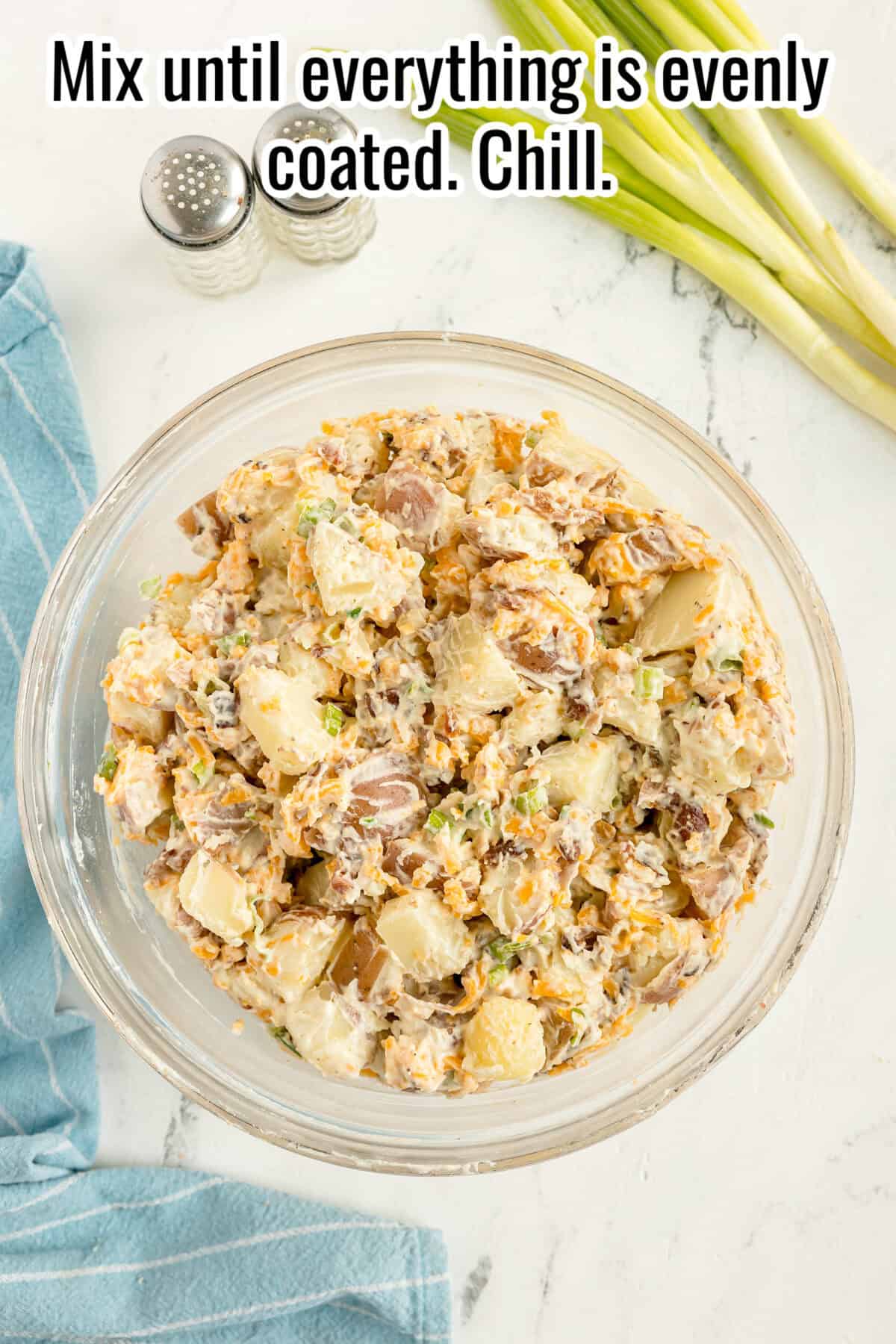 A glass bowl of potato salad mixed with dressing sits on a white table next to green onions, a blue cloth, and salt and pepper shakers. Text above reads, “Mix until everything is evenly coated. Chill.”.