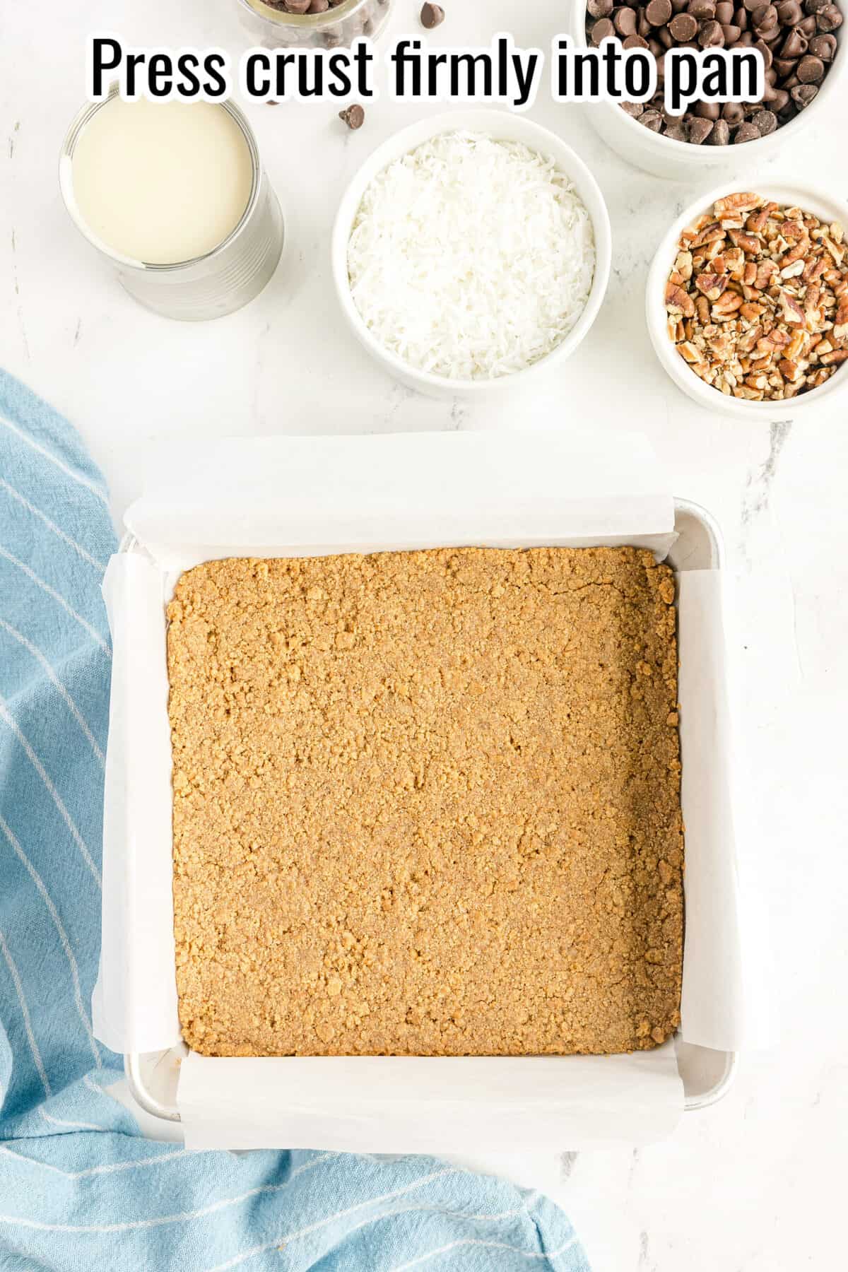 A square baking pan with a pressed crumb crust, surrounded by bowls of sweetened milk, shredded coconut, pecans, and chocolate chips on a white surface.