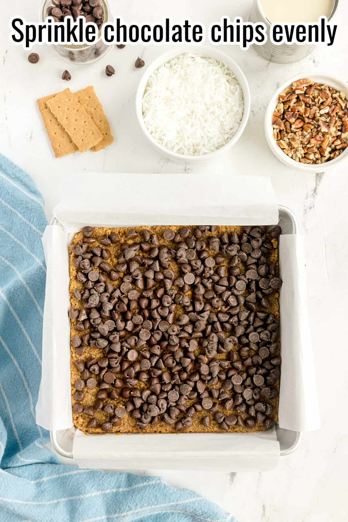A baking pan lined with parchment paper filled with a dessert topped with chocolate chips. Nearby are bowls of coconut, nuts, graham crackers, and a blue towel. Text reads “Sprinkle chocolate chips evenly.”.