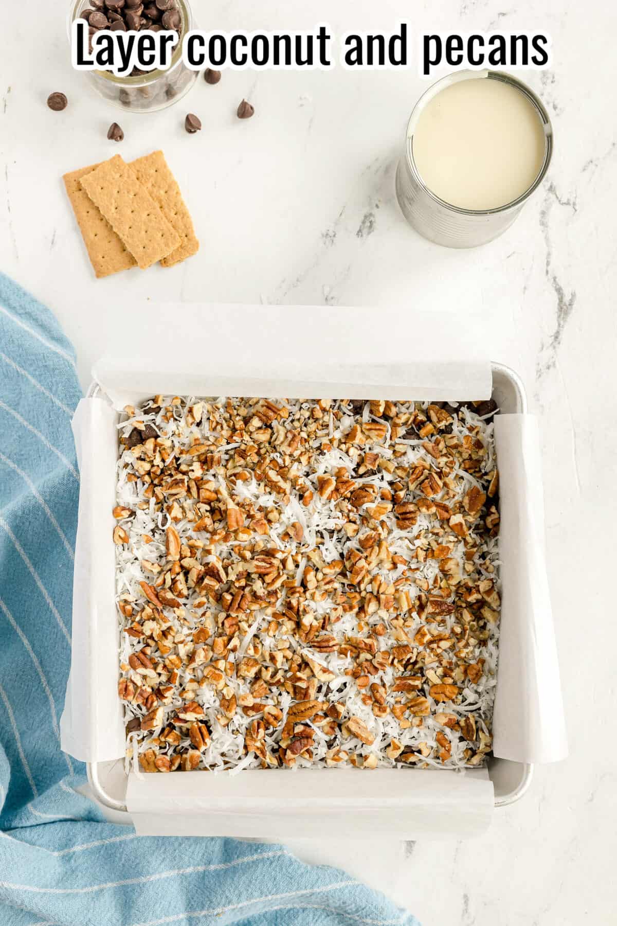 A baking pan lined with parchment paper contains a mixture of coconut and pecans. Nearby are graham crackers, chocolate chips, a can of milk, and a blue towel on a white surface.