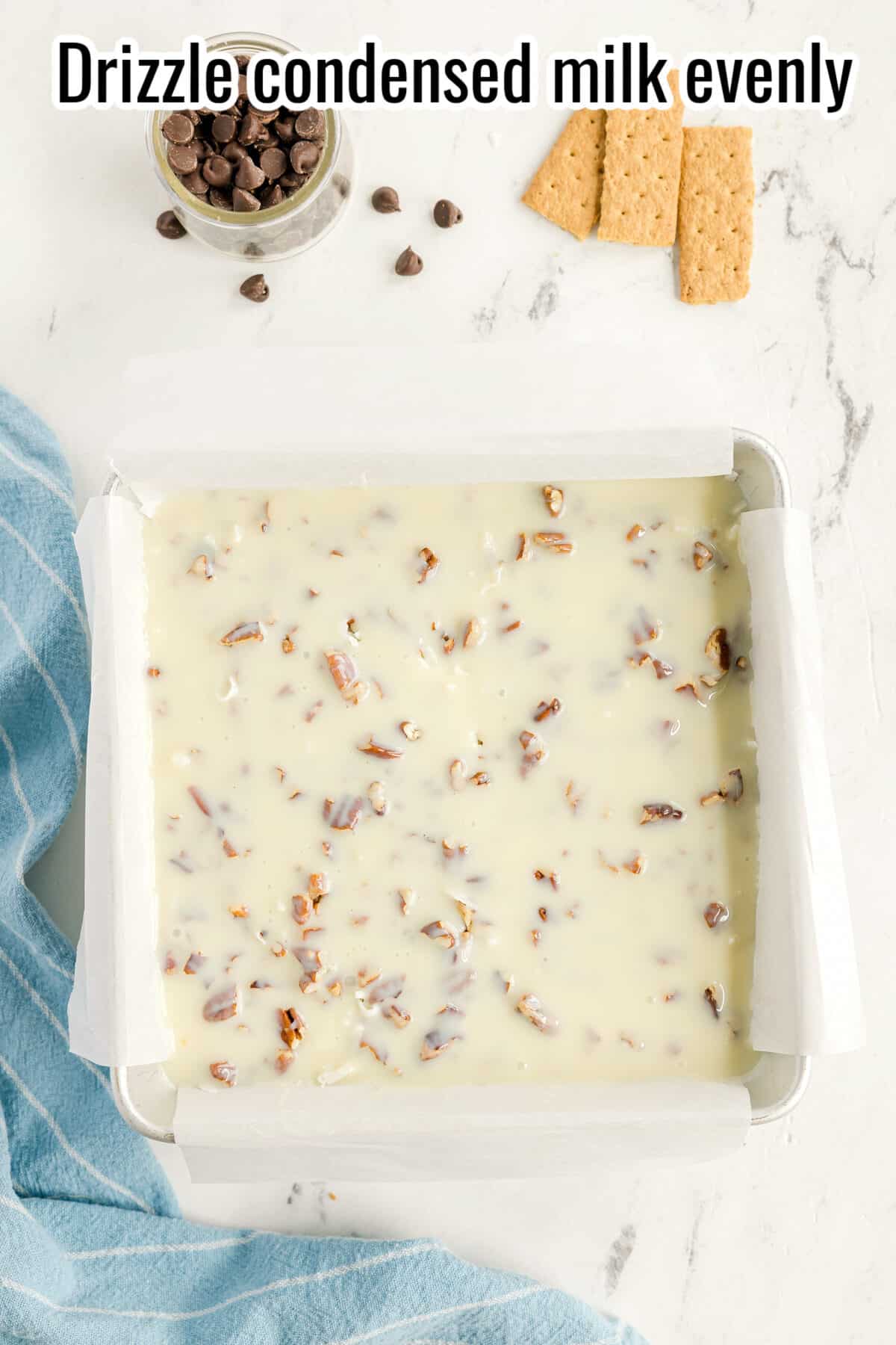 A parchment-lined pan filled with a nut and condensed milk mixture sits on a white surface, with chocolate chips and graham crackers nearby.