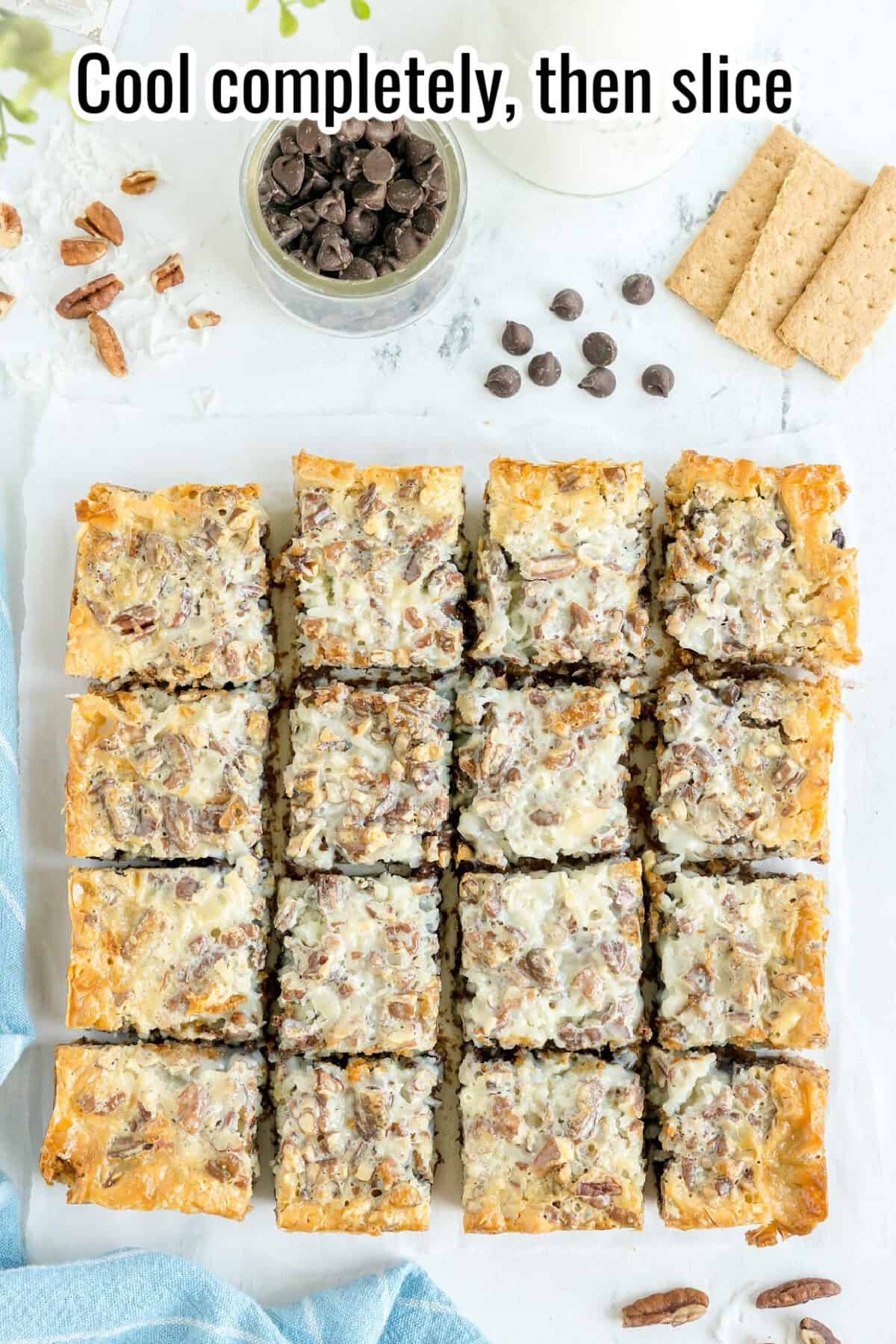Sixteen pecan dessert bars are arranged in a grid on parchment paper, with a bowl of chocolate chips, graham crackers, and chopped pecans nearby. The text reads, “Cool completely, then slice.”.