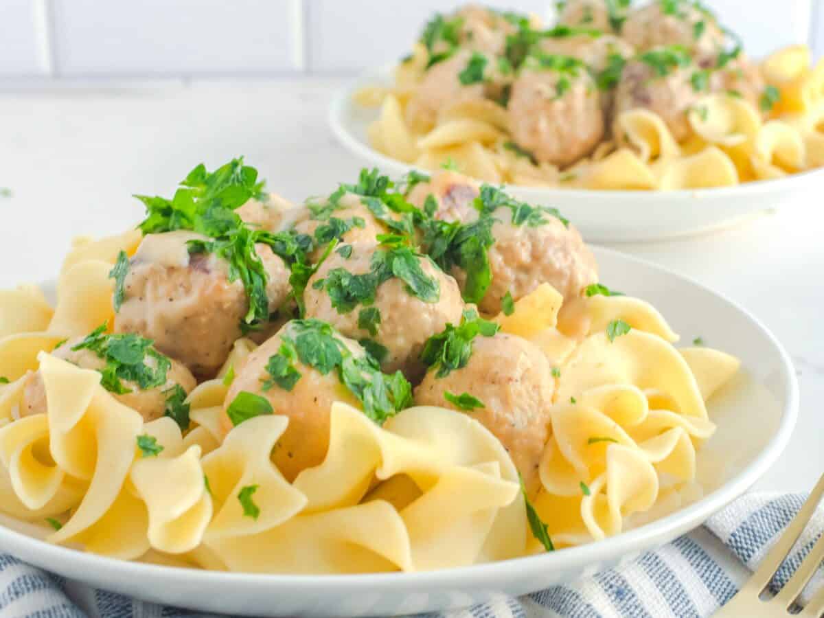 A plate of egg noodles topped with meatballs, creamy sauce, and chopped parsley, with a second plate in the background.