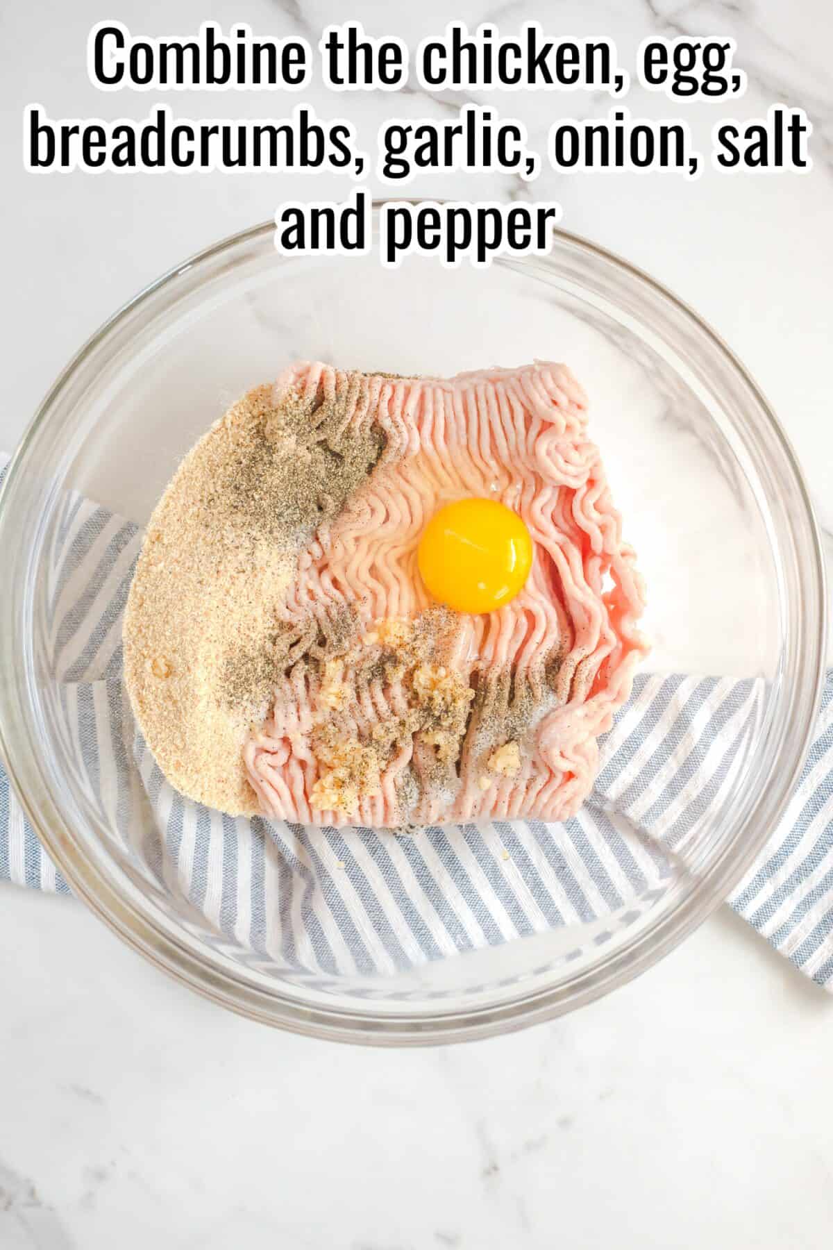 A glass bowl with ground chicken, an egg, breadcrumbs, seasoning, and spices sits on a striped cloth, ready to be mixed.